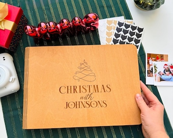 May include: A wooden photo album with the text "Christmas with Johnsons" and a Christmas tree design. The album is on a green and gold striped tablecloth, with a red gift box, a white camera, and a decorative Christmas tree in the background.