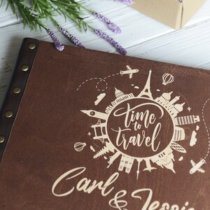 May include: A brown leather photo album with a white inscription that reads "Time to Travel" and "Carl & Jessica". The inscription is surrounded by a circle of iconic landmarks from around the world.