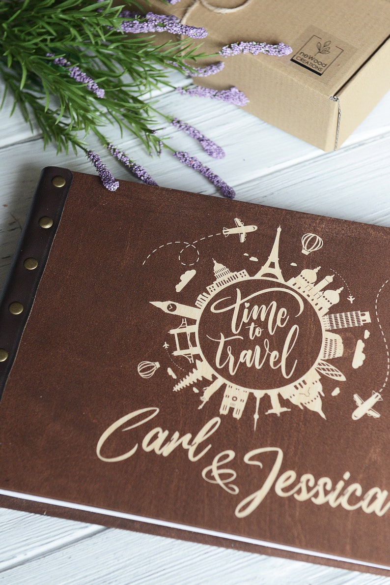 May include: A brown leather photo album with a white inscription that reads "Time to Travel" and "Carl & Jessica". The inscription is surrounded by a circle of iconic landmarks from around the world.