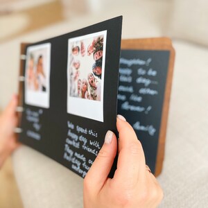 May include: A photo album with a wooden cover and black pages. The album is open, revealing two photos and handwritten text. The photos are of people. The text on the page reads "We spent this happy day with closest friends. They made our holiday fun, unforgettable."