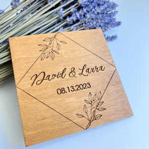 K&ouml;nnte beinhalten: Ein Holzschild in Diamantform mit einem floralen Design. Der Text "David & Laura 08.13.2023" ist in das Schild graviert.