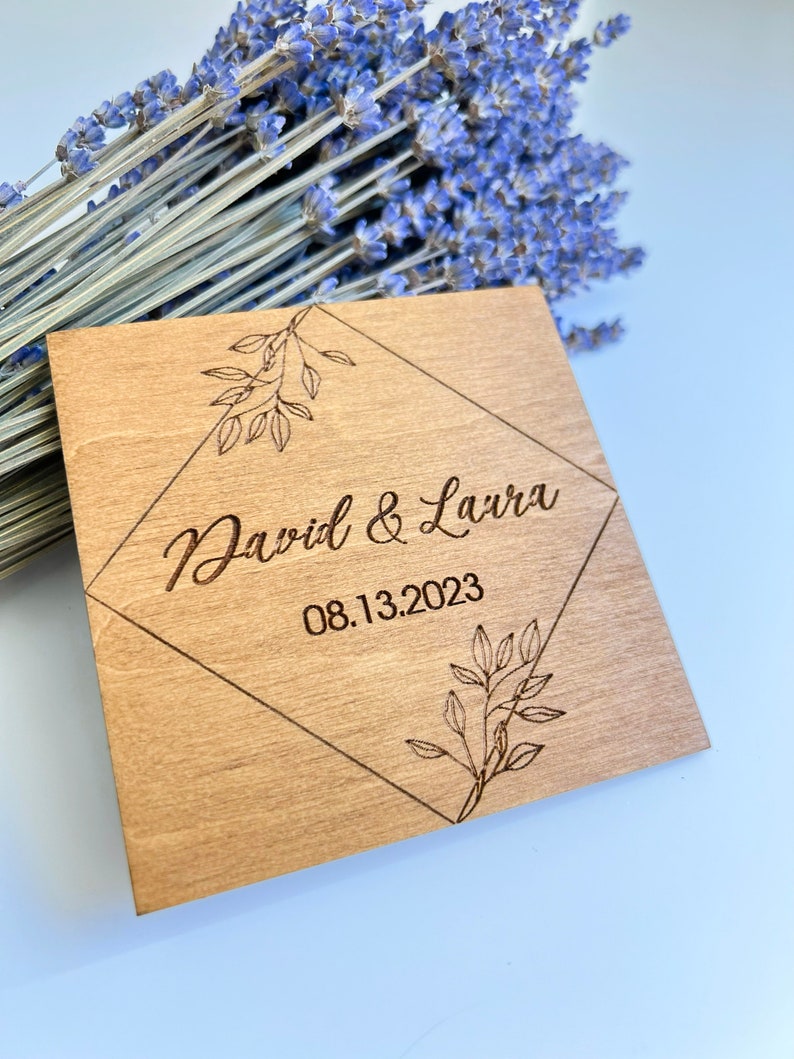 K&ouml;nnte beinhalten: Ein Holzschild in Diamantform mit einem floralen Design. Der Text "David & Laura 08.13.2023" ist in das Schild graviert.