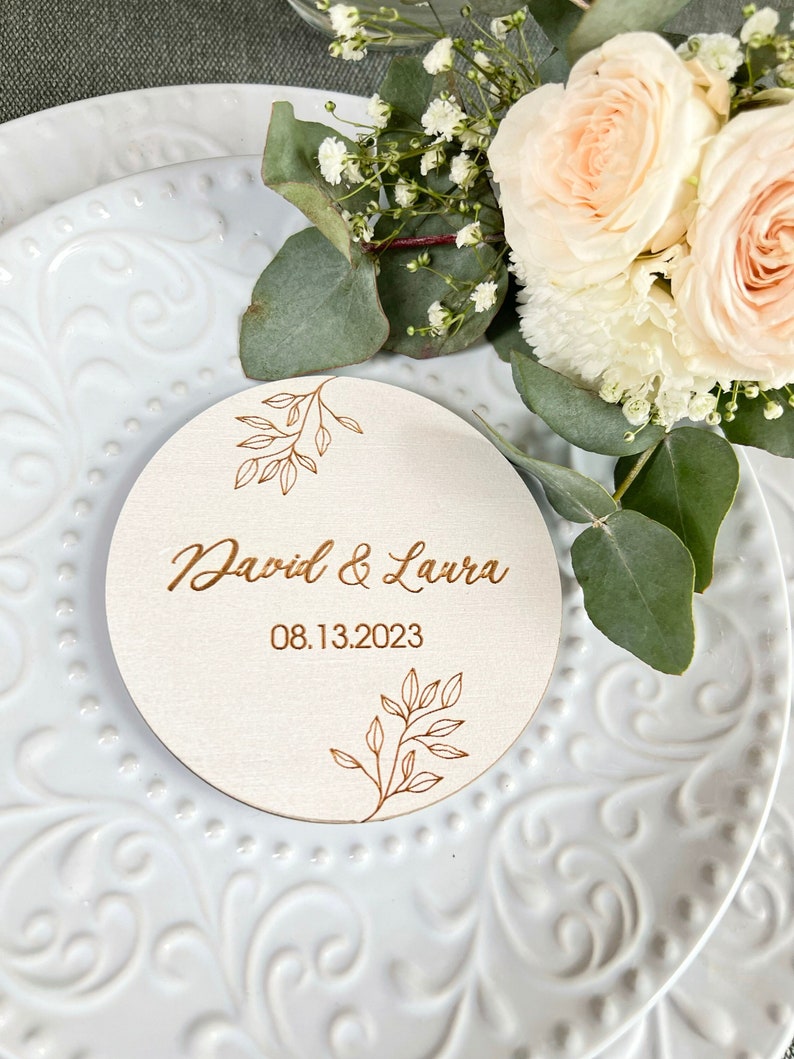 K&ouml;nnte beinhalten: Ein rundes Holzschild mit goldenem Blumendesign und dem Text "David & Laura 08.13.2023" auf wei&szlig;em Hintergrund.