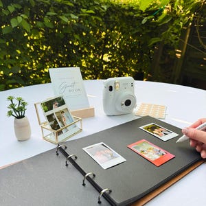 May include: A white instant camera, a photo album, and a gold framed photo holder are arranged on a white surface. A small plant in a beige vase and a "Guest Book" sign are also present. Several instant photos are scattered around the album.