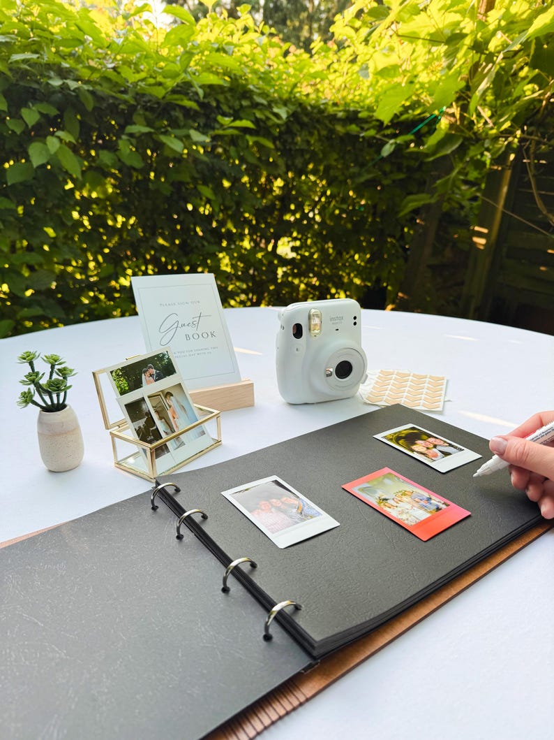 May include: A white instant camera, a photo album, and a gold framed photo holder are arranged on a white surface. A small plant in a beige vase and a "Guest Book" sign are also present. Several instant photos are scattered around the album.