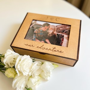 May include: A wooden photo box featuring a photograph of a couple kissing. The box is engraved with the initials "J ♡ C" and the words "our adventure". White roses are arranged on the white surface beside the box.