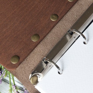 May include: Brown leather binder with a metal ring mechanism and a beige suede backing. The binder has brass rivets along the edge.