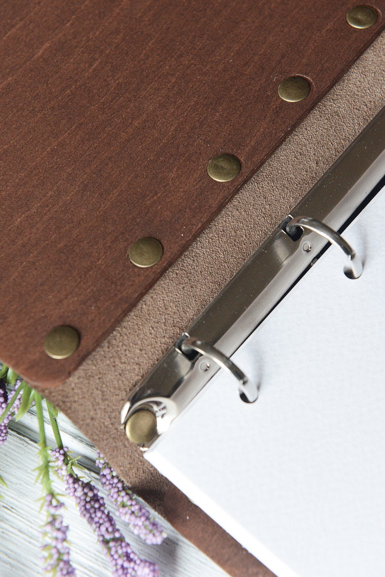 May include: Brown leather binder with a metal ring mechanism and a beige suede backing. The binder has brass rivets along the edge.