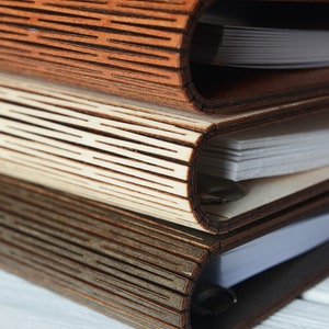 May include: Three wooden journals stacked on top of each other. The covers are made of intricately cut wood, with a textured, layered design. The pages are white and visible within the curved spine of each journal. The wood tones vary from light to dark.
