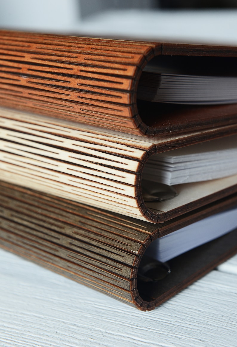 May include: Three wooden journals stacked on top of each other. The covers are made of intricately cut wood, with a textured, layered design. The pages are white and visible within the curved spine of each journal. The wood tones vary from light to dark.