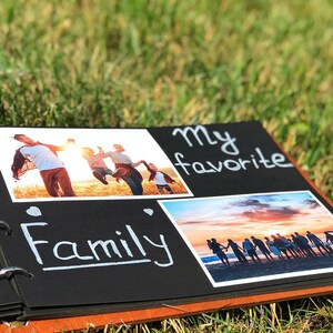 May include: Black photo album with a brown spine. The album has a ring binding and is open to a page with two photos and the text "My favourite Family". The first photo shows a family of four jumping in a field. The second photo shows a group of people standing on a beach at sunset.