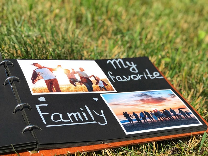 May include: Black photo album with a brown spine. The album has a ring binding and is open to a page with two photos and the text "My favourite Family". The first photo shows a family of four jumping in a field. The second photo shows a group of people standing on a beach at sunset.