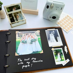 May include: A wedding guest book and photo album with a white Instax camera. The album is open, displaying photos with decorative corner stickers. A sign reads "Guest Book". The album has the text "The best day with my favorites people!"