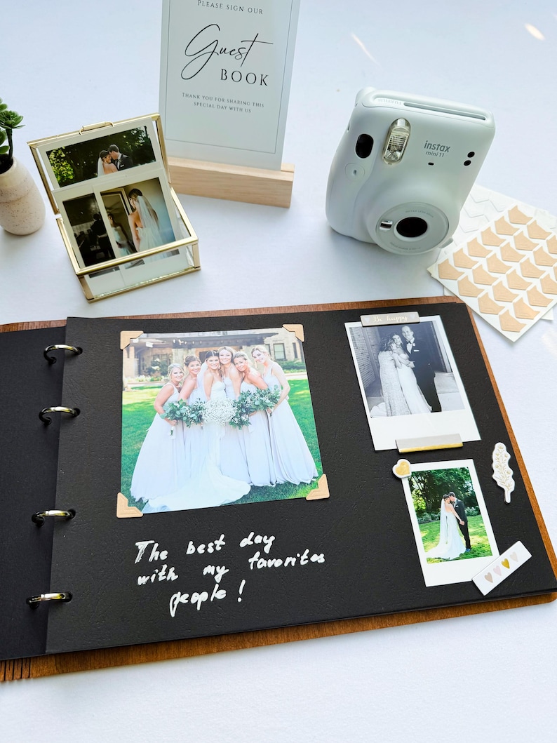 May include: A wedding guest book and photo album with a white Instax camera. The album is open, displaying photos with decorative corner stickers. A sign reads "Guest Book". The album has the text "The best day with my favorites people!"