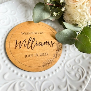 K&ouml;nnte beinhalten: Ein rundes Holzschild mit dem Text "Wedding by Williams July 18, 2023" und einem floralen Design.