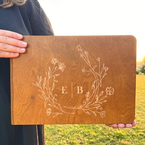 May include: A rectangular wooden sign with a floral wreath design and the initials "E B" in the center. The sign is a warm brown color and is held up in front of a green grassy background.