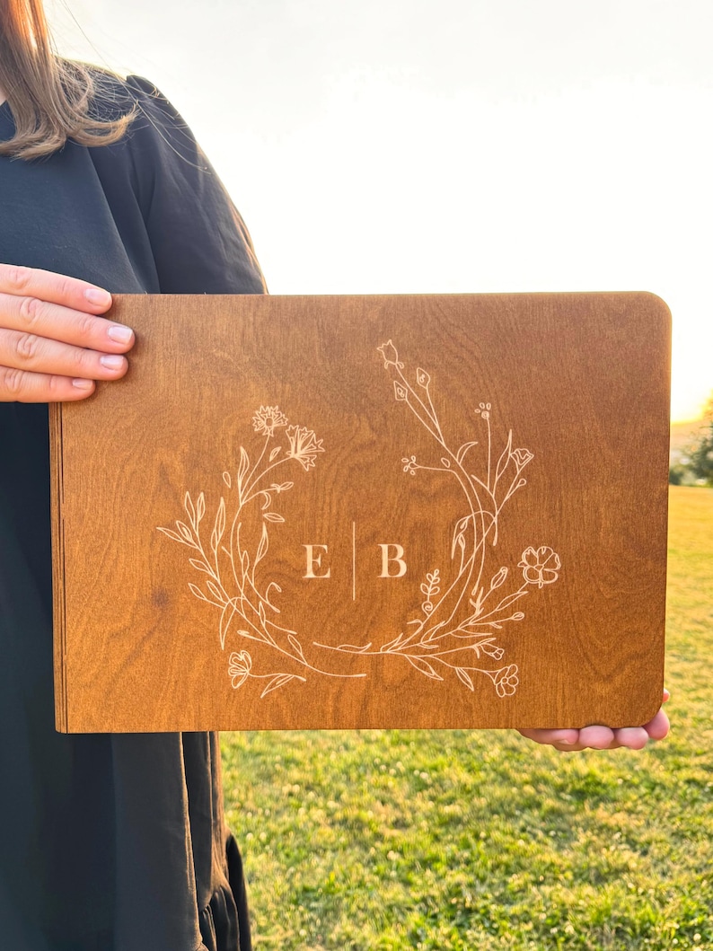 May include: A rectangular wooden sign with a floral wreath design and the initials "E B" in the center. The sign is a warm brown color and is held up in front of a green grassy background.