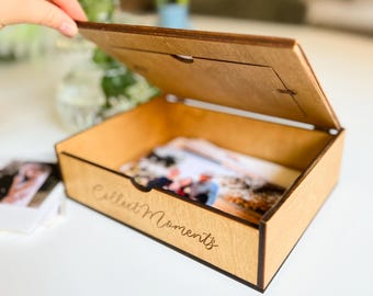 May include: A wooden photo box with a hinged lid and a cutout for a photo. The box is light brown and has the words "Collect Moments" engraved on the front.