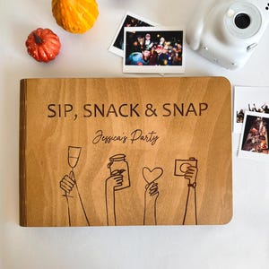 Op de afbeelding: Een houten gastenboek met de woorden "SIP, SNACK & SNAP" en "Jessica's Party" gegraveerd op de kaft. De kaft heeft een lijntekening van handen die een champagneglas, pot, hart en camera vasthouden. Een witte instant camera en foto's zijn ook aanwezig.