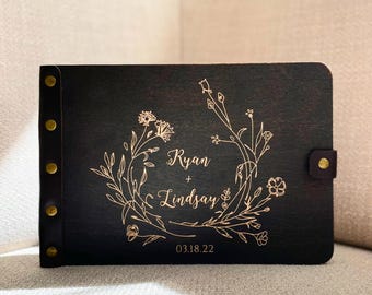 Op de afbeelding: Zwart houten fotoalbum met een gouden bloemenkrans ontwerp en de namen "Ryan + Lindsay" gegraveerd. De datum "03.18.22" is ook aanwezig. Met leren riem sluiting en messing klinknagels. Lengte ongeveer 20 cm.
