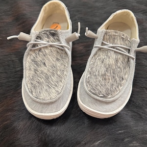 May include: A pair of gray slip-on shoes with a cowhide design on the top. The shoes have white soles and gray laces. The shoes are displayed on a dark, textured surface.