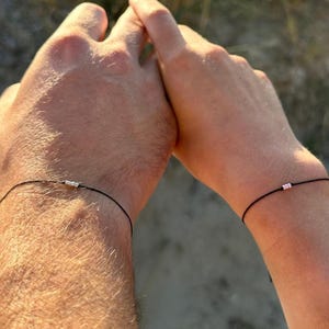 Könnte beinhalten: Zwei dünne schwarze Kordelarmbänder an den Handgelenken. Ein Armband hat eine kleine Reihe silberfarbener Metallperlen, während das andere Armband eine kleine Reihe hellrosa Perlen aufweist. Die Armbänder sind schlicht, minimalistisch und zart.
