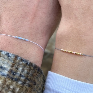 Op de afbeelding: Twee bijpassende vriendschapsarmbanden met kleine kralen. De ene armband is wit met lichtblauwe kralen en de andere is grijs met roze en gouden kralen.