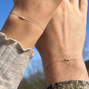 Könnte beinhalten: Zwei zarte Armbänder mit winzigen silbernen Perlen auf einer durchsichtigen Schnur. Die Armbänder werden an den Handgelenken von zwei Personen getragen.
