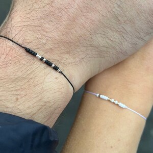 Könnte beinhalten: Zwei Armbänder mit Perlen. Ein Armband hat schwarze und silberne Perlen auf einer schwarzen Kordel. Das andere Armband hat weiße und goldene Perlen auf einer hellvioletten Kordel. Die Armbänder werden an den Handgelenken getragen.