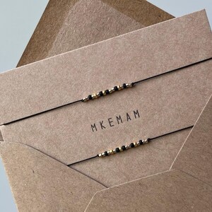 Può includere: Due braccialetti di corda nera con perline dorate su una busta marrone. La busta ha la scritta "MKEMAM" stampata.