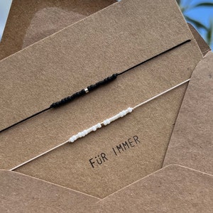 Può includere: Due braccialetti dell'amicizia su una carta marrone con il testo "Für Immer", che significa "Per sempre" in tedesco. I braccialetti sono fatti con perline nere e bianche.