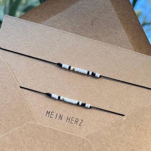 Könnte beinhalten: Zwei schwarze Armbänder aus Schnur mit weißen und schwarzen Perlen und einem goldenen Perlenakzent. Die Armbänder befinden sich auf einem braunen Papierumschlag mit dem Text "MEIN HERZ" darauf.