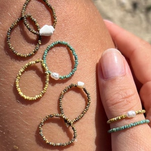 Könnte beinhalten: Ein Set aus sechs Perlenringen mit Perlenakzenten. Die Ringe sind aus kleinen, bunten Perlen in Braun-, Gelb- und Türkistönen gefertigt. Einige der Ringe haben einen einzelnen Perlenakzent, während andere zwei haben.