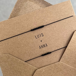 Könnte beinhalten: Zwei schwarze Armbänder mit Perlenakzenten werden auf einer braunen Karte in einem braunen Umschlag gezeigt. Auf der Karte steht "LUIS + ANNA" in schwarzer Schrift. Die Armbänder sind minimalistisch.