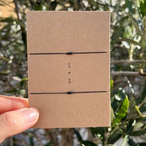 May include: Two black string bracelets on a brown card. The card has the letters "S + S" printed on it. The background is out of focus, with green leaves and branches.