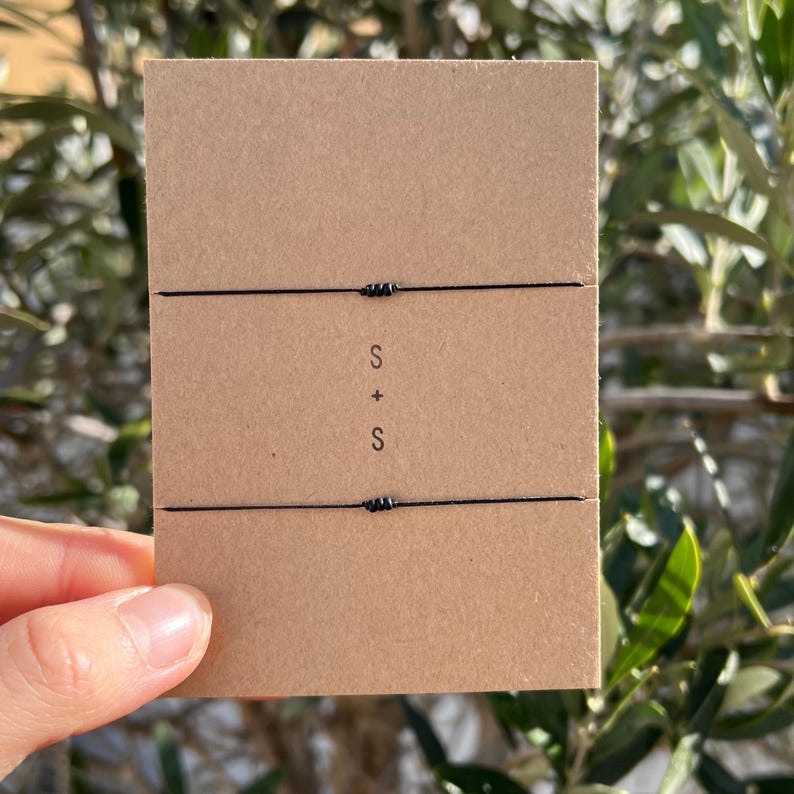 May include: Two black string bracelets on a brown card. The card has the letters "S + S" printed on it. The background is out of focus, with green leaves and branches.