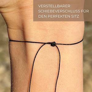 May include: A black adjustable cord bracelet with a sliding knot closure. The bracelet is worn on a person's wrist. The text "VERSTELLBARER SCHIEBEVERSCHLUSS F&Uuml;R DEN PERFEKTEN SITZ" is visible above the bracelet.
