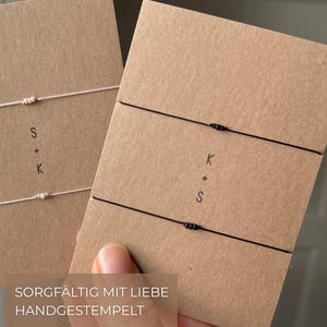 May include: Two handmade bracelets on brown cards. One bracelet has a white cord with a knot and the letters "S + K". The other bracelet has a black cord with a knot and the letters "K + S". The text "Sorgf&auml;ltig mit Liebe handgestempelt" is at the bottom.