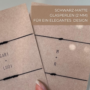 May include: Two kraft paper cards with black string bracelets. One card has the names "SARI + LUDI" printed on it. The other card has the initials "M + R". The text "SCHWARZ-MATTE GLASPERLEN (2 MM) F&Uuml;R EIN ELEGANTES DESIGN" is printed on a brown rectangle.