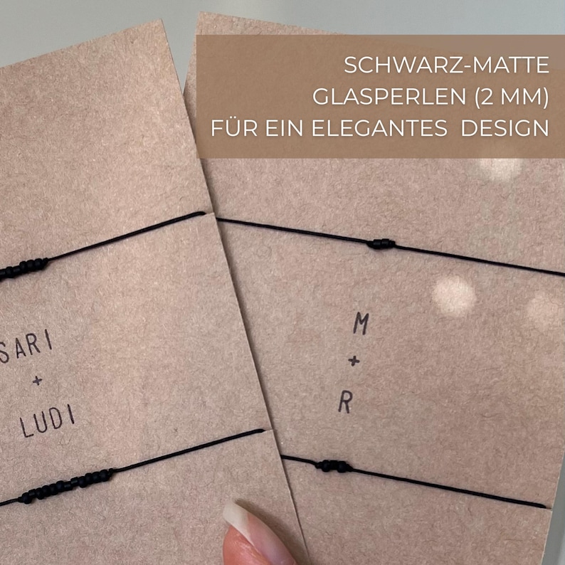 May include: Two kraft paper cards with black string bracelets. One card has the names "SARI + LUDI" printed on it. The other card has the initials "M + R". The text "SCHWARZ-MATTE GLASPERLEN (2 MM) F&Uuml;R EIN ELEGANTES DESIGN" is printed on a brown rectangle.