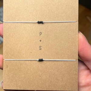 May include: Two matching bracelets with black beads on a thin, light-coloured cord. The bracelets are displayed on a brown card with the letters "P + S" printed in the centre. The background is a neutral colour.