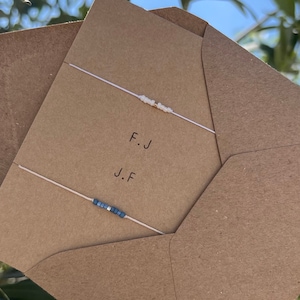 May include: Two friendship bracelets on a brown envelope. The top bracelet is white with gold beads and the bottom bracelet is blue. The envelope has the initials "F.J" and "J.F" written on it.