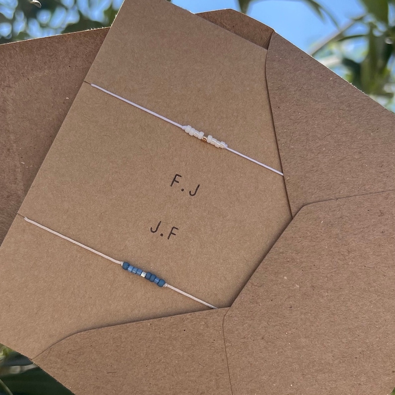May include: Two friendship bracelets on a brown envelope. The top bracelet is white with gold beads and the bottom bracelet is blue. The envelope has the initials "F.J" and "J.F" written on it.
