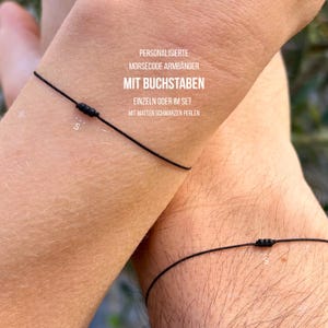 Può includere: Primo piano di due polsi, ognuno adornato con un braccialetto di filo nero con perline nere opache. I braccialetti hanno un design minimalista. Il testo nell'immagine recita: "PERSONALISIERTE MORSECODE ARMBÄNDER MIT BUCHSTABEN EINZELN ODER IM SET MIT MATTEN SCHWARZEN PERLEN."