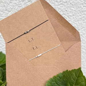 May include: A brown paper envelope with two friendship bracelets inside. The bracelets are made with black and white beads and string. The envelope has the initials "L.T." and "T.L." written on it.