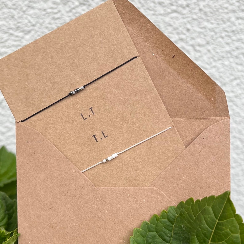 May include: A brown paper envelope with two friendship bracelets inside. The bracelets are made with black and white beads and string. The envelope has the initials "L.T." and "T.L." written on it.