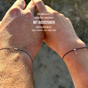 Könnte beinhalten: Zwei schwarze Armbänder mit kleinen silbernen und rosa Perlen. Ein Armband hat eine silberne Perle, das andere hat drei rosa Perlen und den Buchstaben "S". Der Text in deutscher Sprache ist ebenfalls sichtbar.
