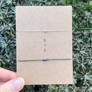 Könnte beinhalten: Zwei Freundschaftsarmbänder auf einer braunen Karte. Das obere Armband ist weiß mit rosa und grünen Perlen. Das untere Armband ist schwarz mit blauen Perlen. Die Karte zeigt "G + C".