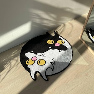Könnte beinhalten: Ein runder Teppich mit einem Cartoon-Katzen-Design. Der Teppich ist in zwei Hälften geteilt, eine schwarze und eine weiße, wobei sich die Gesichter der Katzen überlappen. Die Katzen haben gelbe Augen und rosa Nasen. Der Teppich liegt auf einem Holzboden.