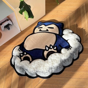 May include: A cartoon character rug in blue and tan, set on a white cloud-shaped base. The rug is placed on a wooden floor. A gaming console and a plant are visible in the background.